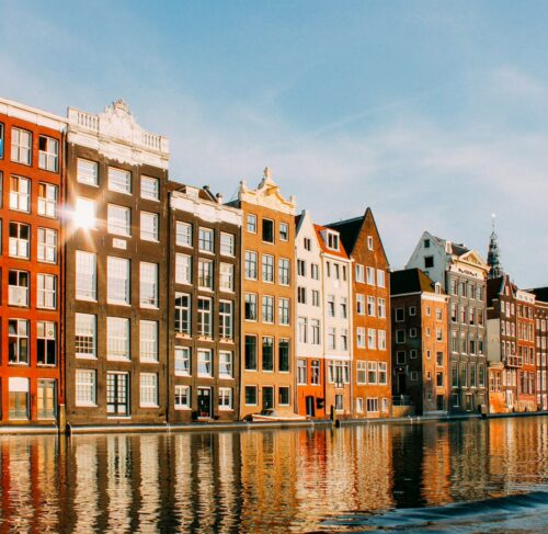 Row of colorful historic canal houses reflected in Amsterdam's calm canal water with trees lining the waterfront