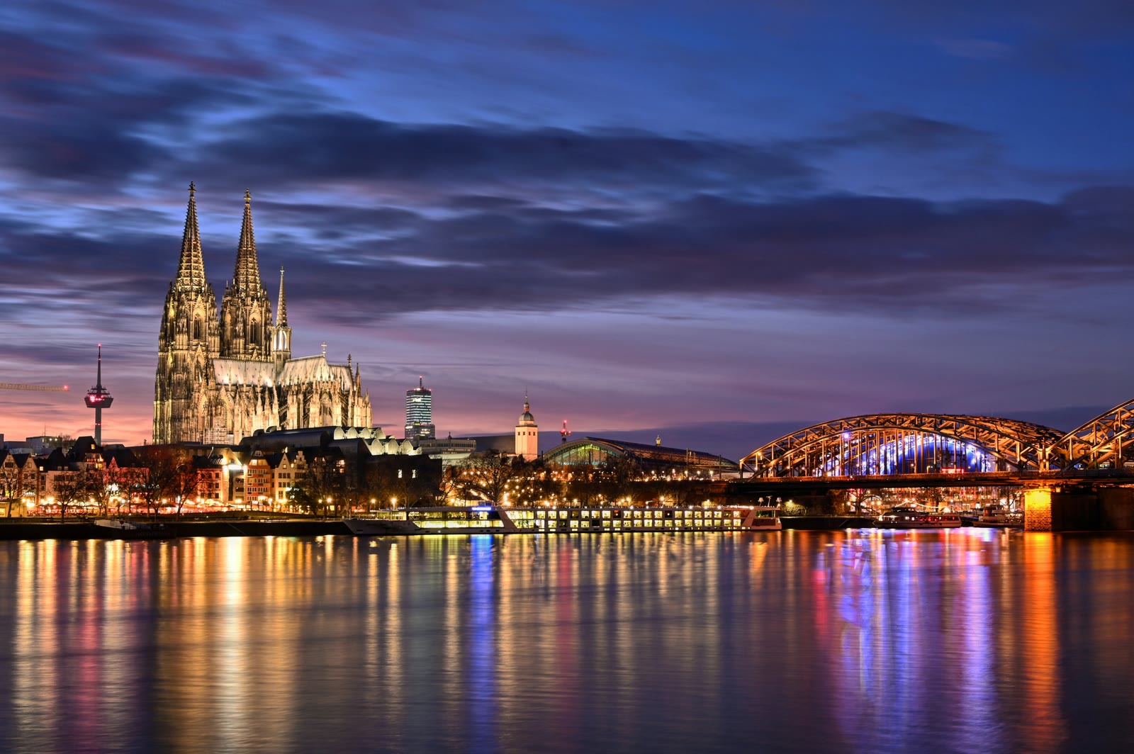 Cologne Cathedral and Hohenzollern Bridge illuminated at night across the Rhine River
