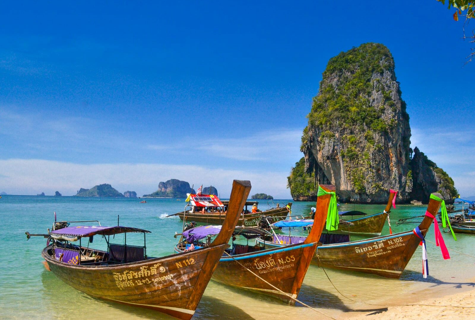 Five traditional wooden longtail boats moored on emerald water with limestone karst cliffs in background, Krabi Thailand