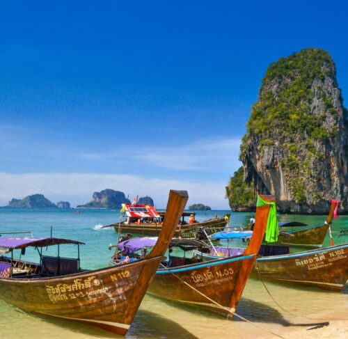 Five traditional wooden longtail boats moored on emerald water with limestone karst cliffs in background, Krabi Thailand