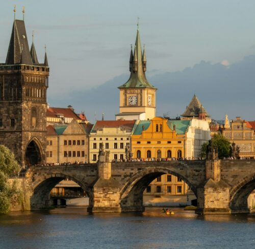View of Charles Bridge spanning the Vltava River with Prague's Old Town buildings and spires in the background under soft daylight